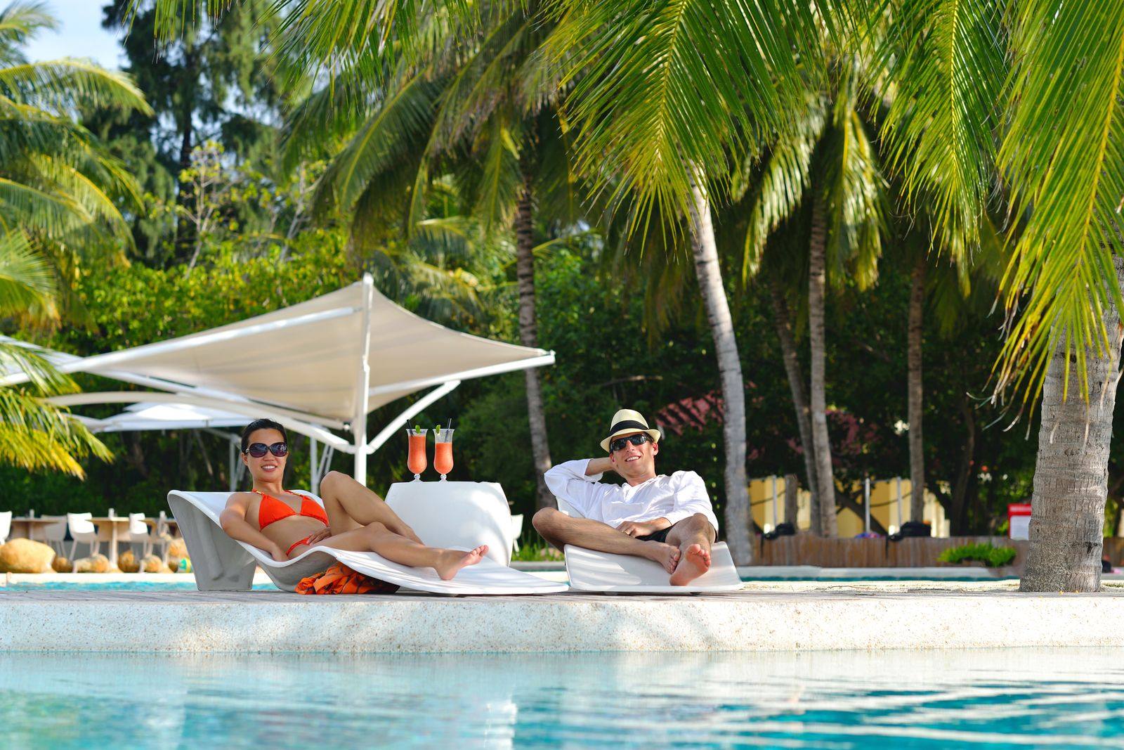 Couple in sunglasses relaxing on white poolside loungers with drinks, under shade sails and palm trees