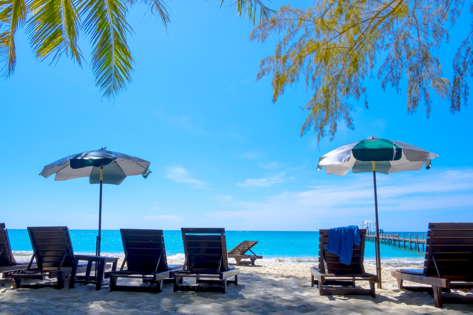 Beach chairs and umbrellas on white sand facing bright turquoise water, with palm fronds overhead and a small pier in the distance