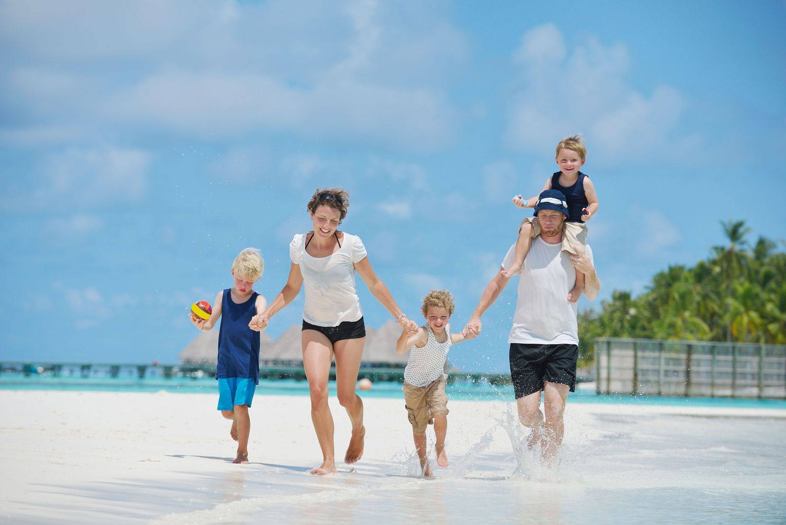 Parents and three young children running hand in hand along a white-sand beach, one child on dad's shoulders