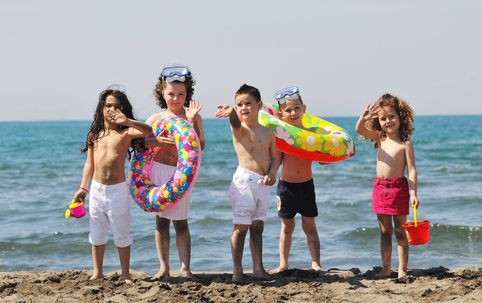 Five children in swimsuits holding pool floats, a snorkel mask and a sand bucket at the edge of the sea