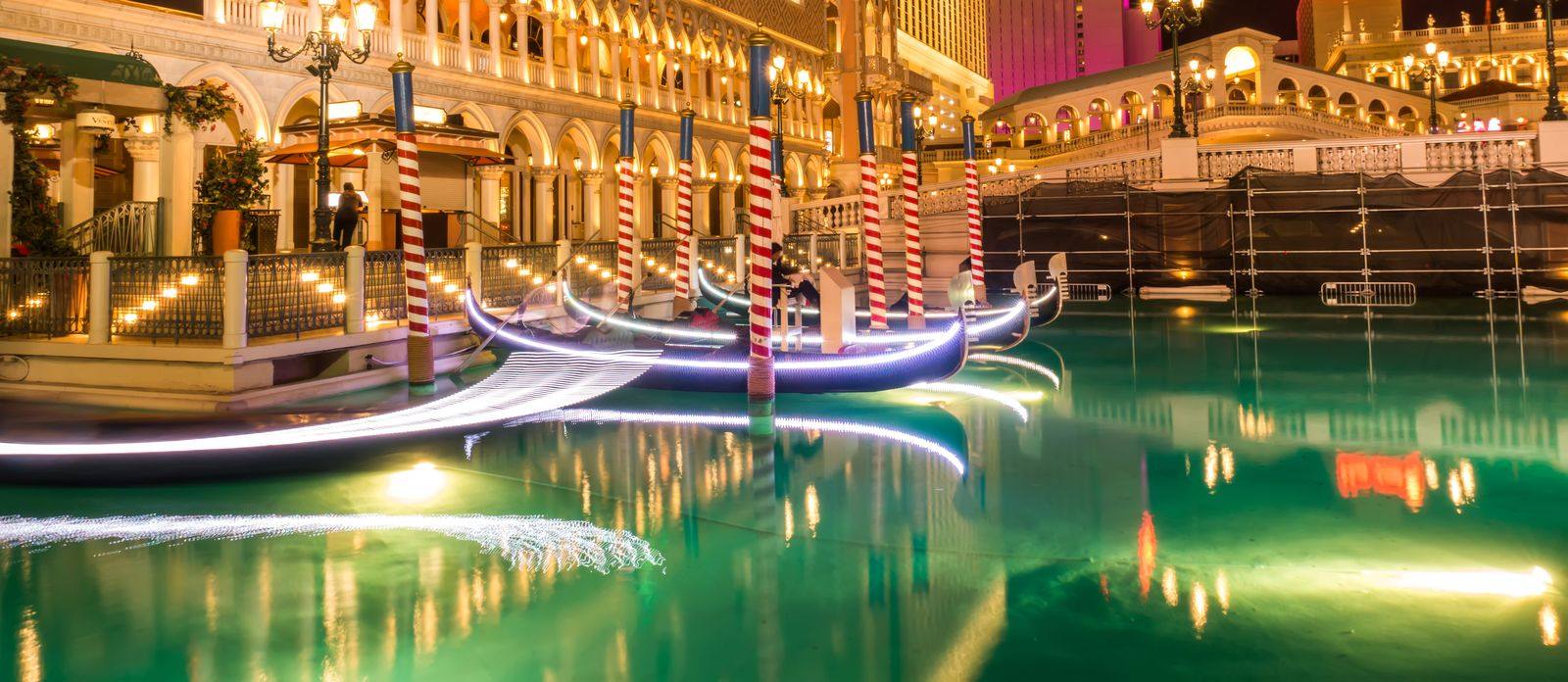 Gondolas with striped mooring poles on a lit canal beside arched Venetian-style buildings at night in Las Vegas
