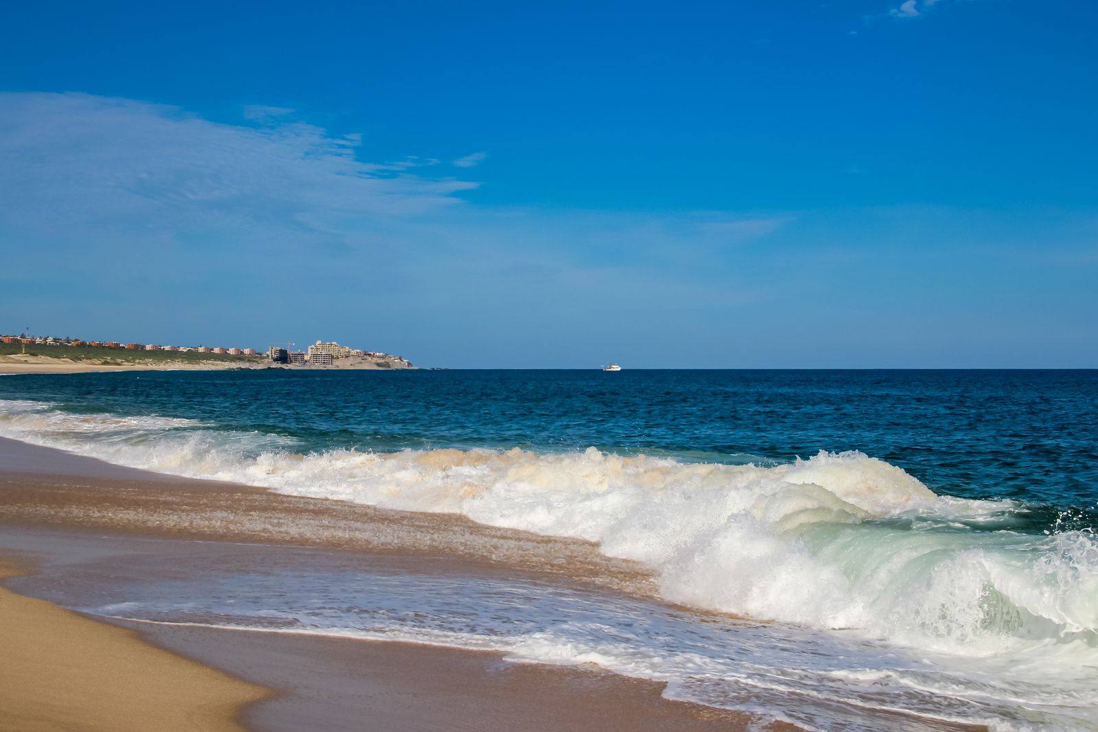 Waves curling onto a wide sandy beach at Cabo San Lucas, with a low headland and buildings in the distance under a clear blue sky