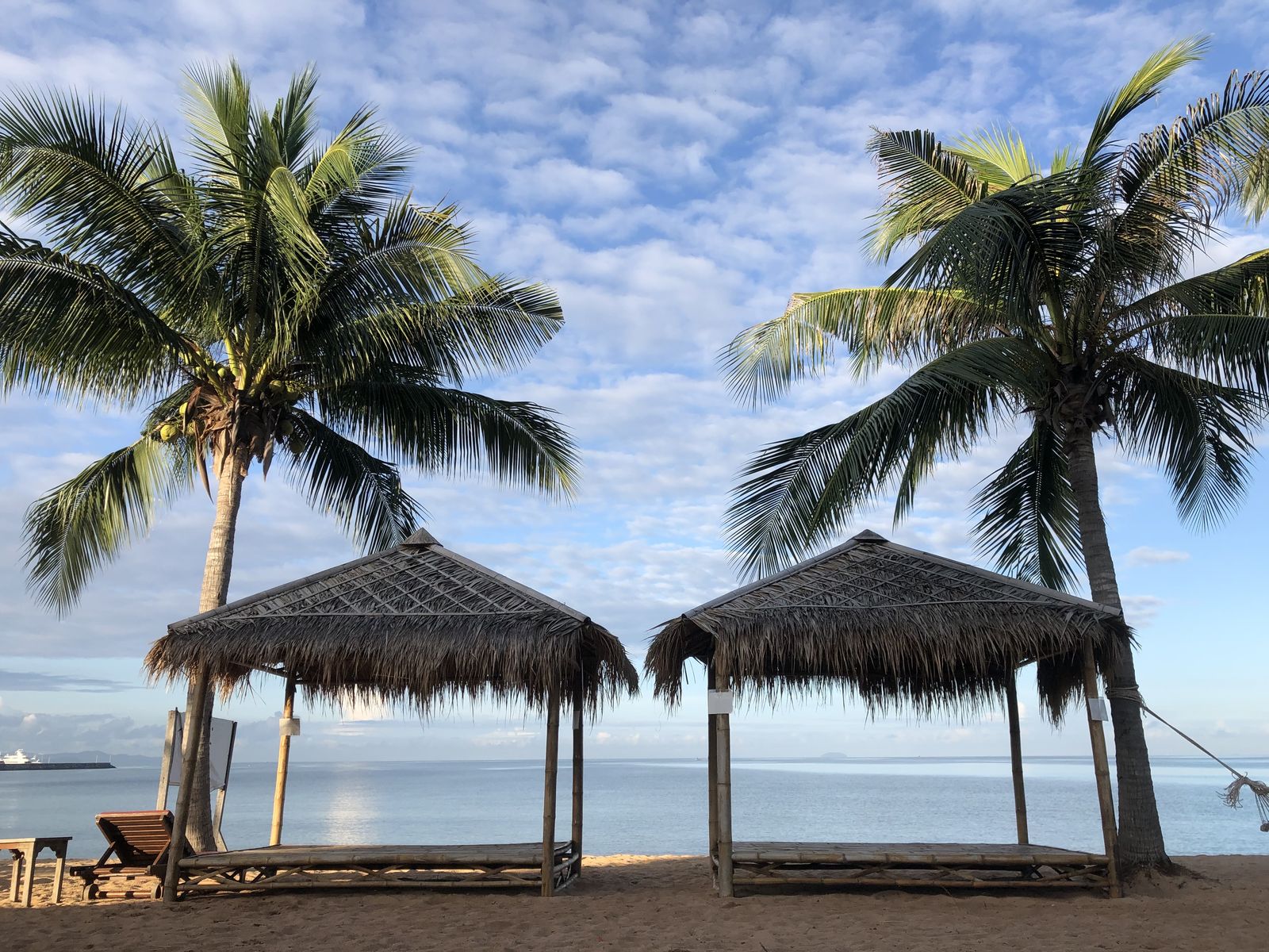 Two thatched-roof shelters on a sandy beach between tall palm trees under a blue sky