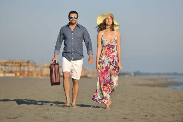 Couple walking barefoot on a sandy beach, the man carrying a small suitcase