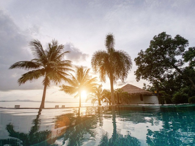 Infinity pool at a tropical resort at sunset, with palm trees and a thatched building reflected in the water