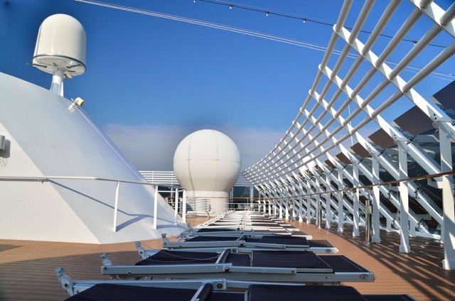 Sun deck of a cruise ship with lounge chairs under white overhead beams and two radar domes, against a blue sky