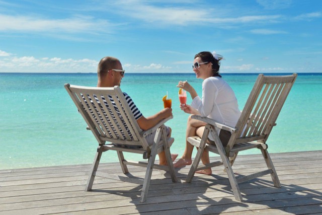 Couple sitting in wooden chairs on a beach deck with drinks, turquoise water behind them