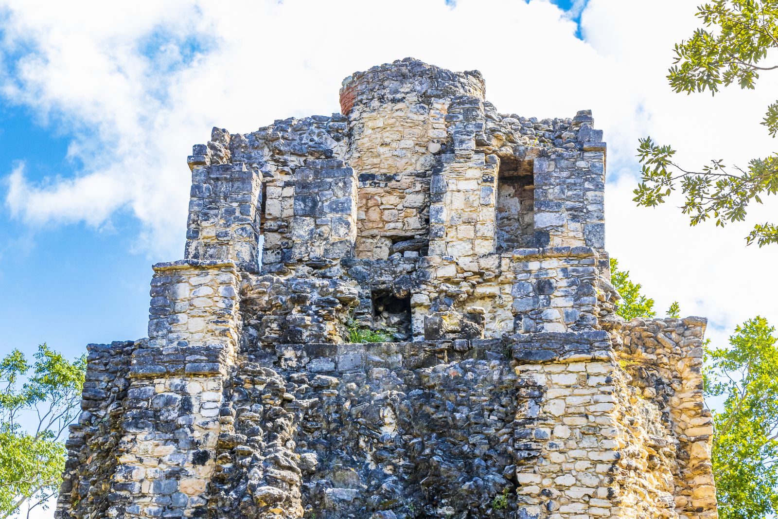 Weathered stone Mayan temple rising above green trees under a partly cloudy sky at Muyil, Mexico