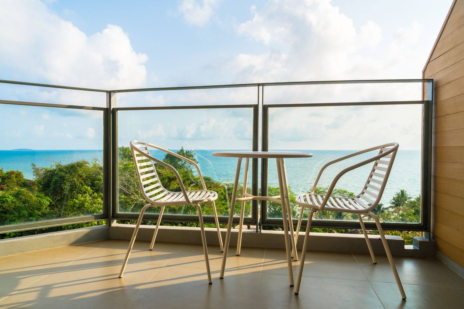 Balcony with two white chairs and a small table behind a glass railing, looking out over treetops to the sea