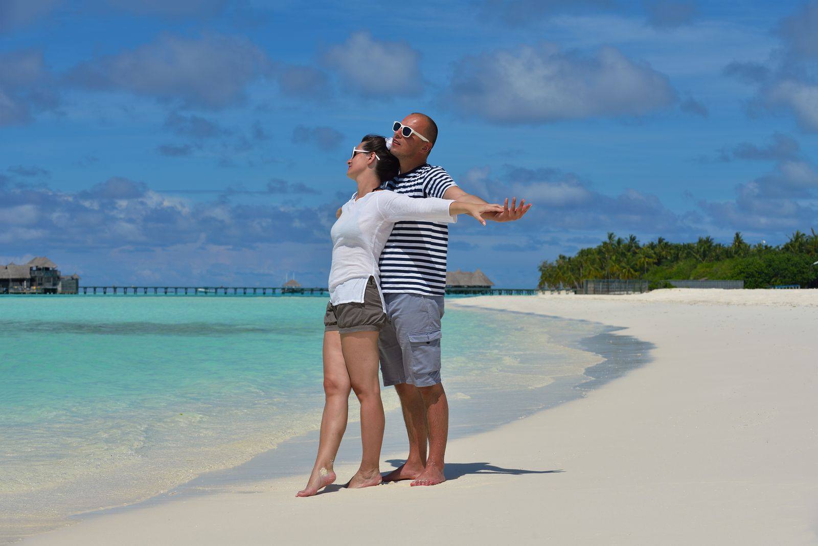 Couple standing on a white sand beach beside turquoise water, one with arms spread wide