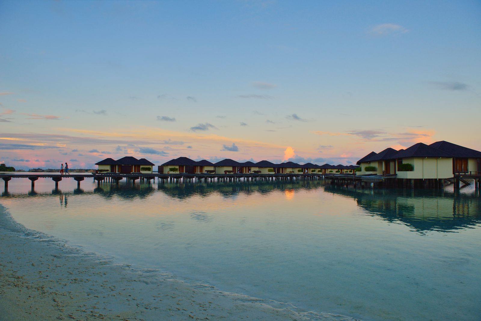 Row of overwater villas joined by a long wooden walkway, reflected in calm water at dusk