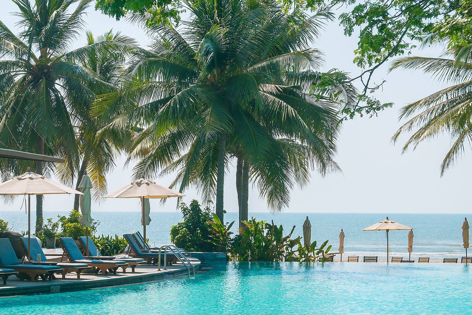 Resort pool with blue loungers and white umbrellas under palm trees, with the sea behind