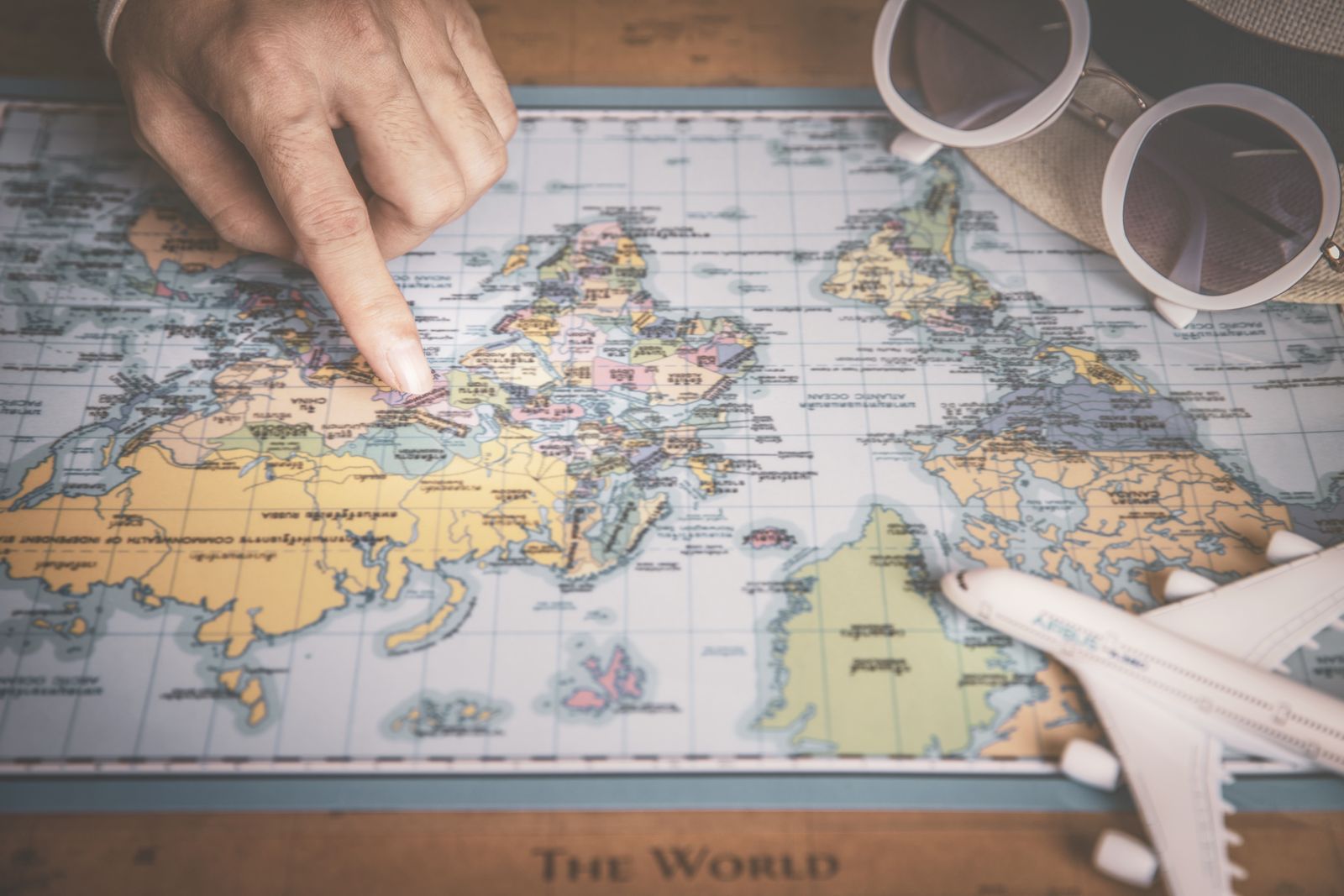 A hand pointing at a world map with a toy airplane and sunglasses beside it
